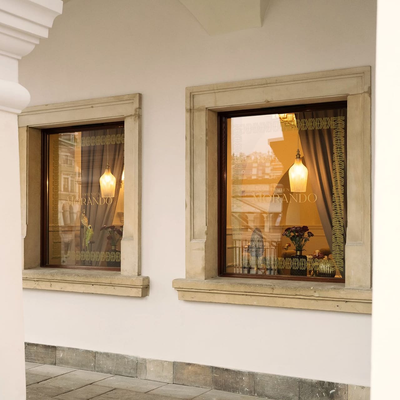 The Kamienica Morando arcade: two windows in stone frames between white pillars, “Morando” on the glass, warm lamplight inside