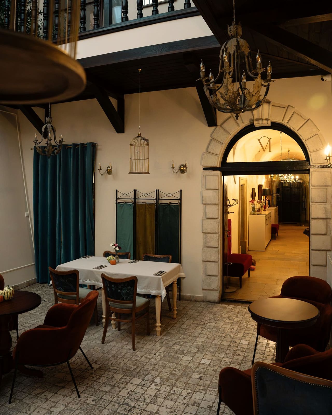The courtyard of the house: laid tables and velvet armchairs, wrought-iron chandeliers and a stone arched doorway with an “M” monogram leading to a lit corridor