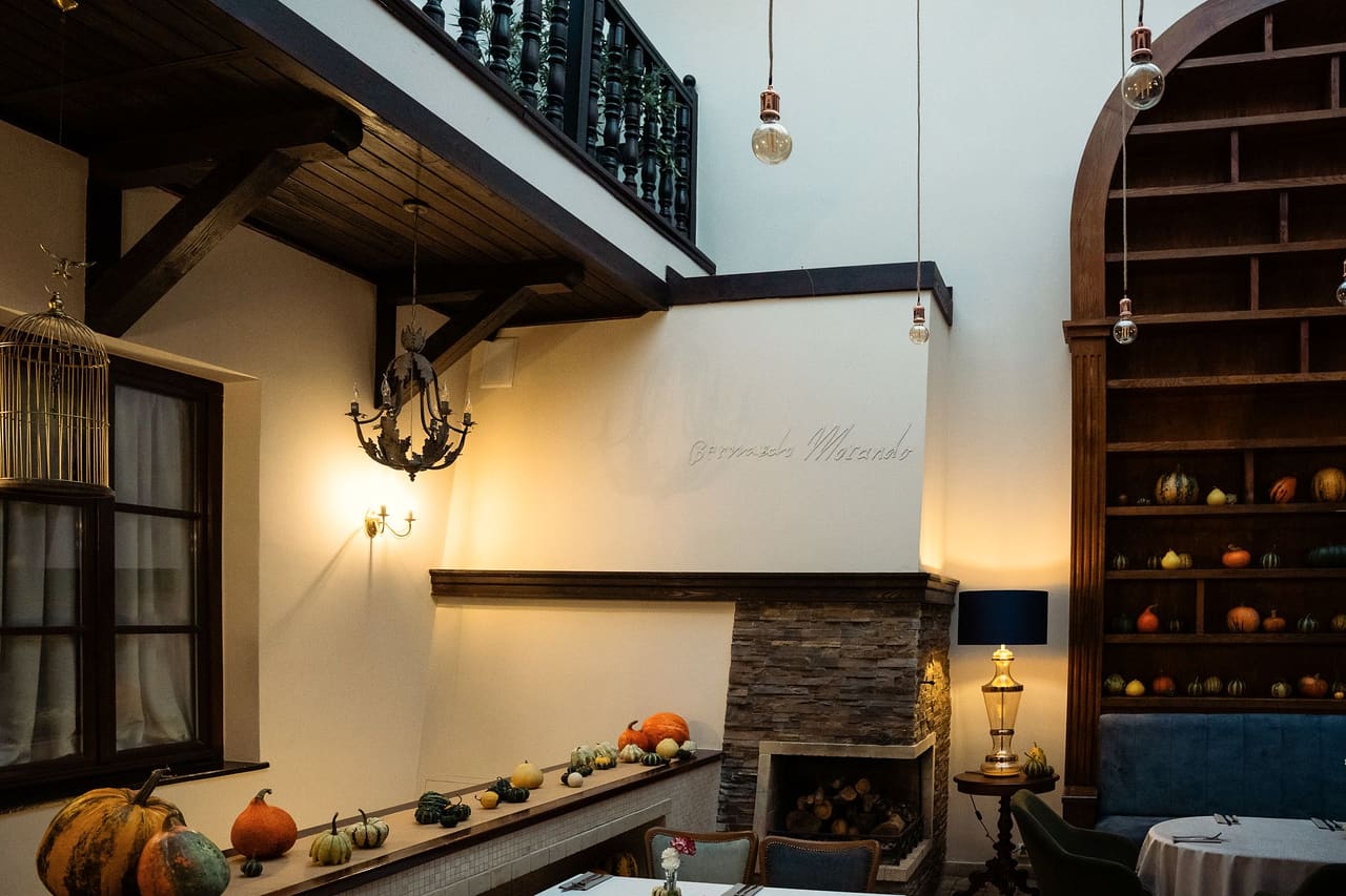 The courtyard of the house: a wall lettered “Bernardo Morando” above the stone fireplace, a row of squashes on the ledge, the wooden gallery with its balustrade above, a shelf unit full of squashes beside it