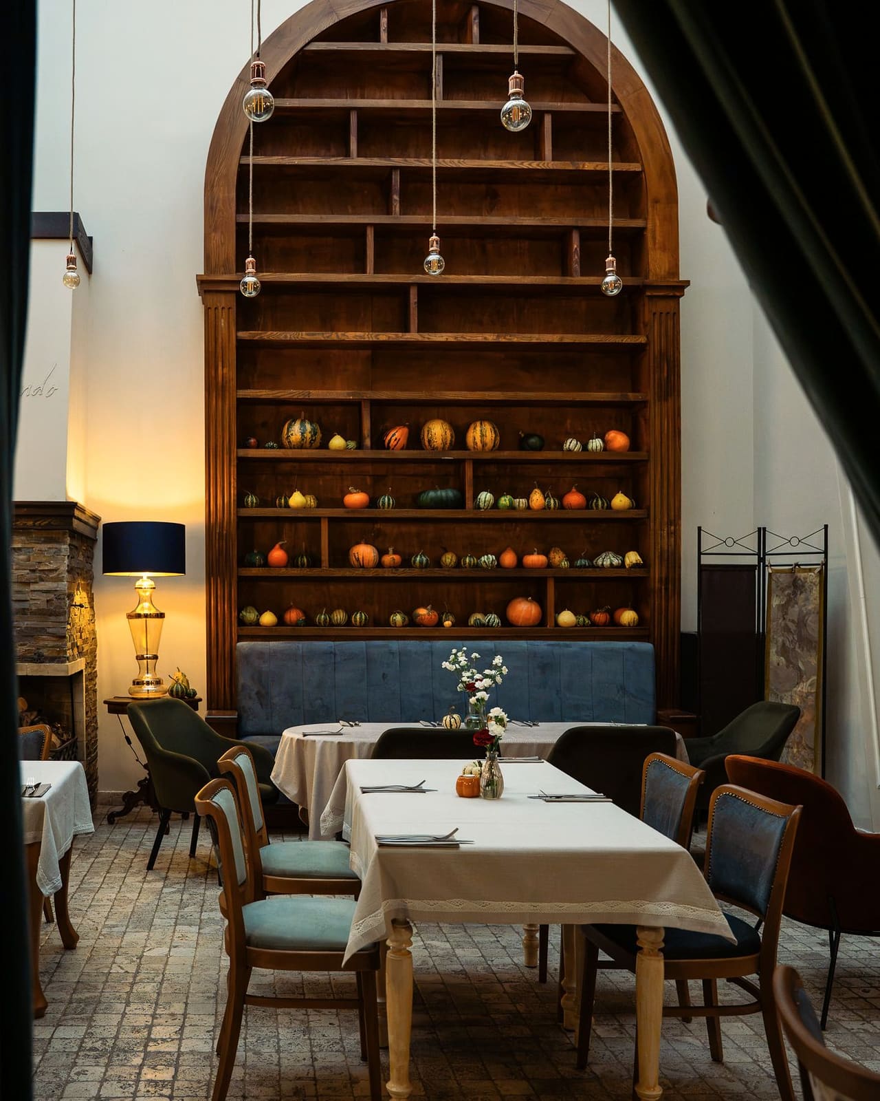 A long table with a white cloth in the courtyard, chairs with blue upholstery around it, an arched shelf unit with squashes, a lamp and a navy banquette beyond