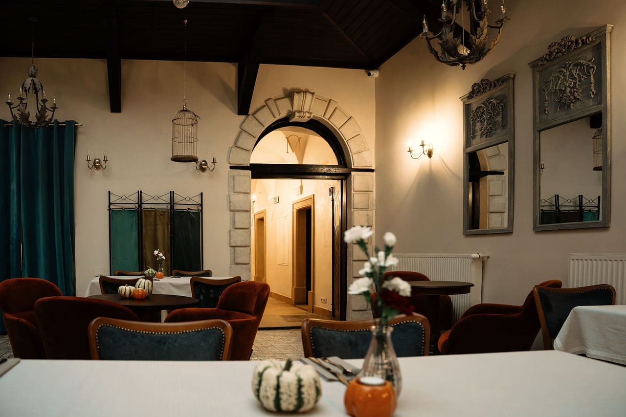 A laid table with white carnations, a small squash and a candle holder in the courtyard, burgundy armchairs, mirrors in ornate frames and a stone arched doorway behind