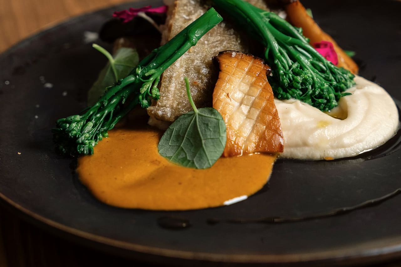 Cod in crayfish sauce with celeriac purée, oyster mushroom and wild broccoli on a dark plate, a wine glass behind on the wooden table