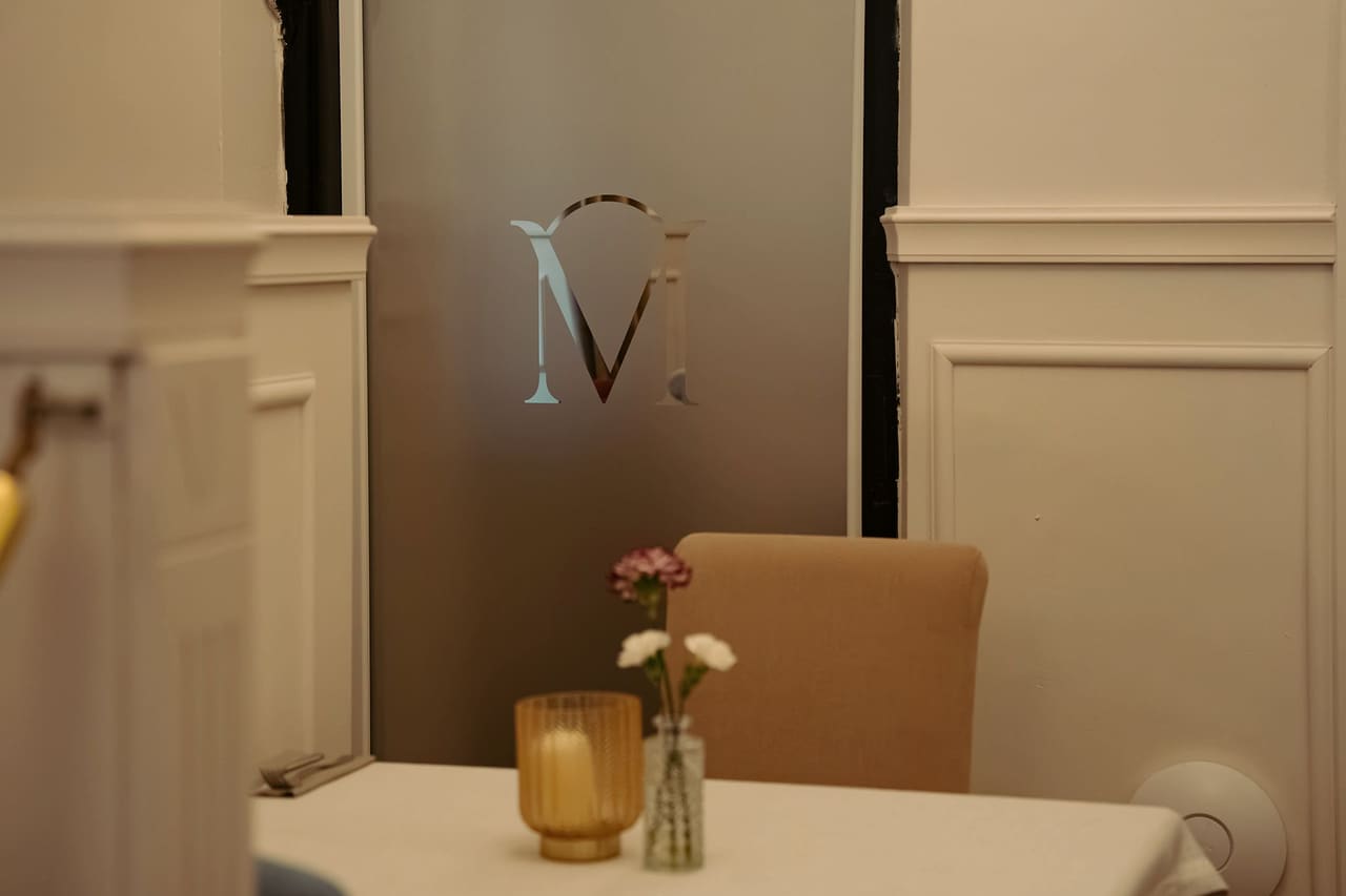 A frosted glass door with an “M” monogram, in front of it a small table with carnations in a vase and a candle in a golden holder, white panelling