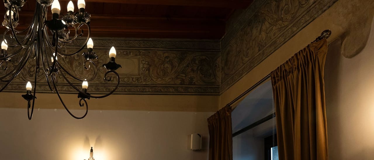 A painted frieze of plant ornament below the wooden ceiling of a room, a wrought-iron candle chandelier in front of it, a golden curtain at the window