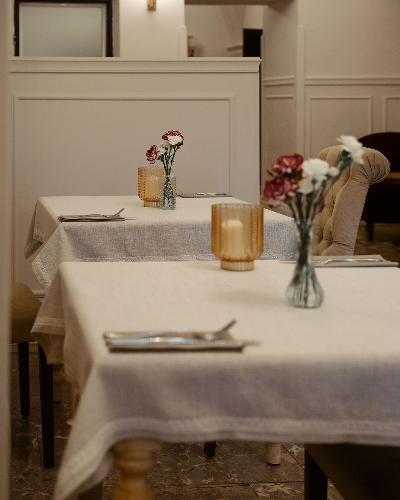 A row of tables laid with linen cloths in a room with white panelling: cutlery on napkins, carnations in vases, candles in golden holders