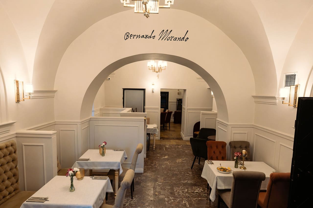 Sala restauracji pod sklepieniem: nad łukiem przejścia napis „Bernardo Morando”, żyrandol, stoliki nakryte białymi obrusami, z kwiatami w wazonikach