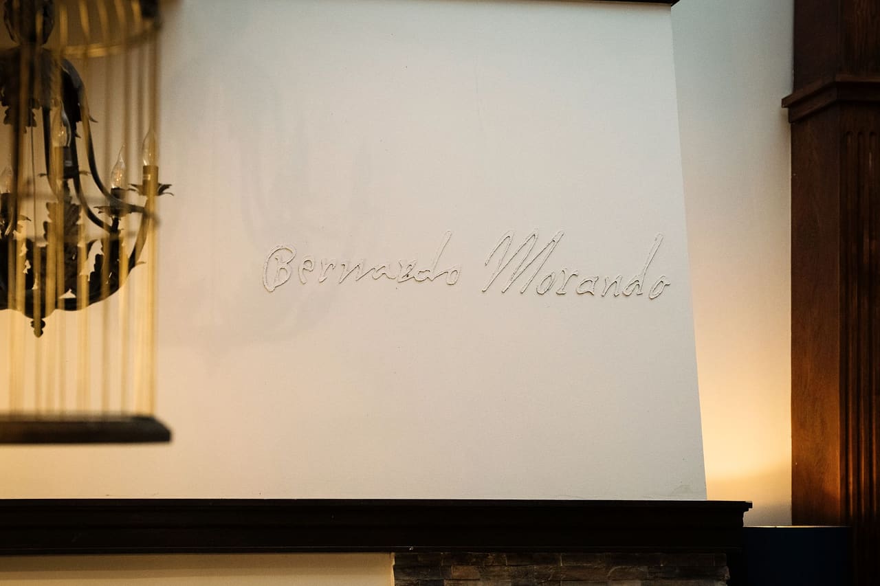 “Bernardo Morando” in raised letters on the white wall above the stone fireplace, a blurred golden chandelier on the left